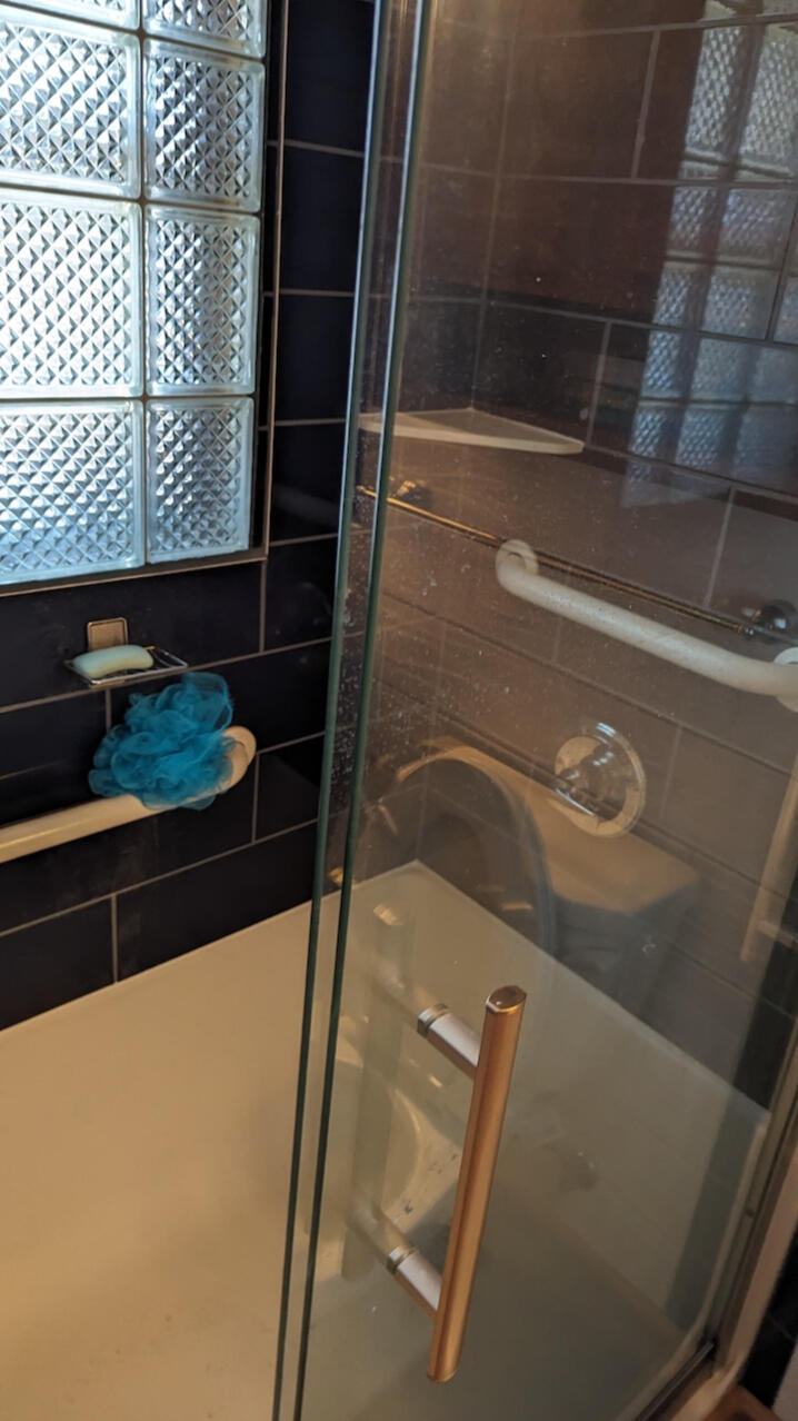 Shower Remodel - Third Day Plumbing Glass shower door with a metal handle, a blue bath pouf hanging on a grab bar, and tiled walls surrounding a bathtub—expertly installed by Third Day Plumbing - Des Moines Iowa.