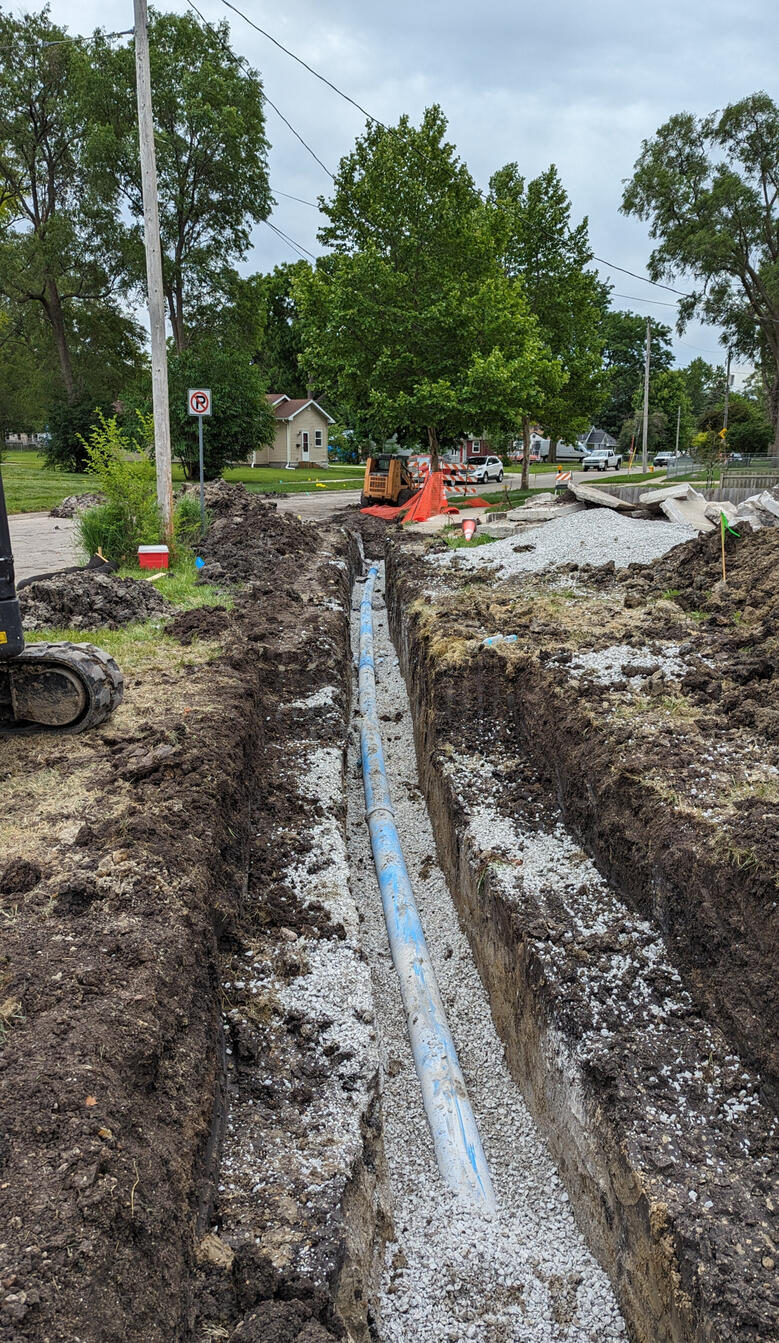Water pipes dug in a trench - Third Day Plumbing - Des Moines Iowa A blue water pipe lies in a freshly dug trench along a roadside construction site, with gravel and construction equipment visible in the background.